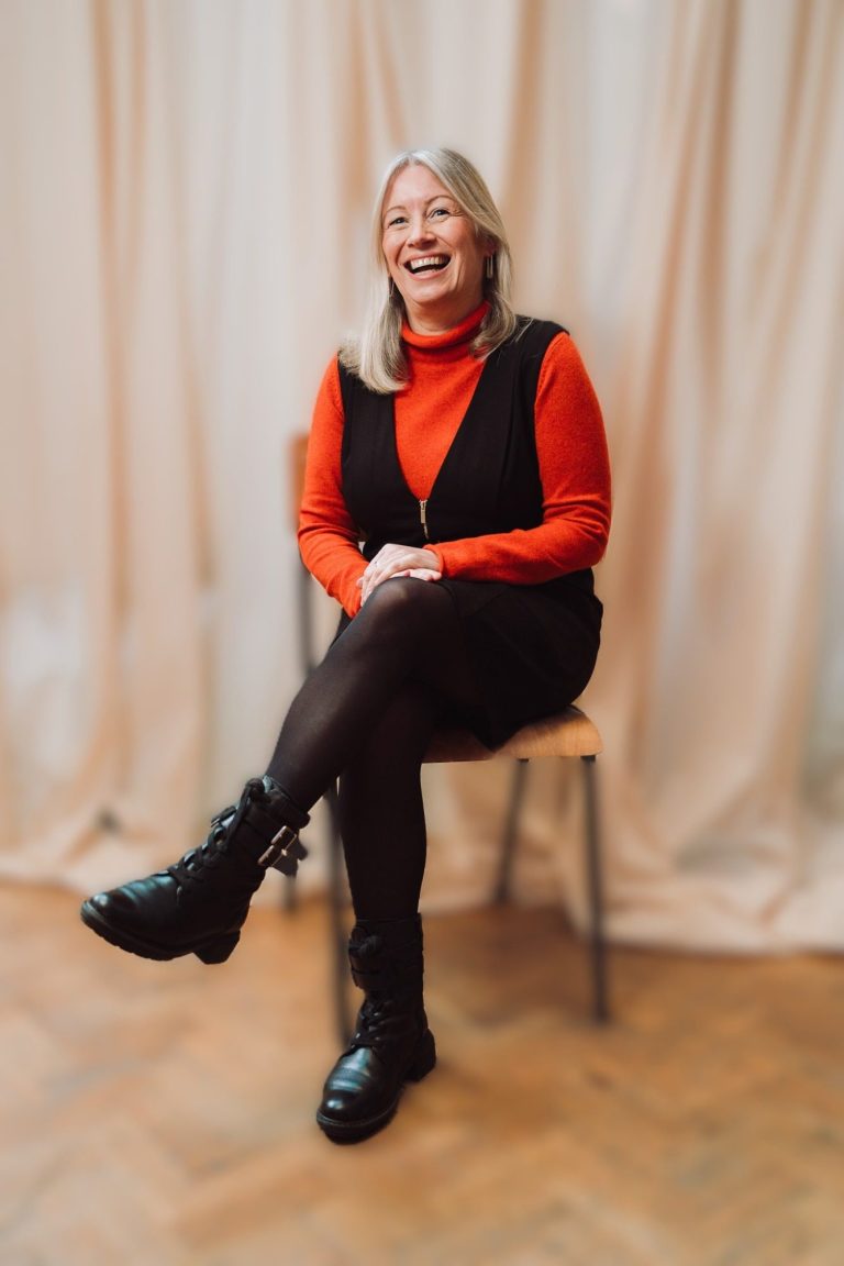 Woman with long hair in a red top and black pinafore dress, sitting on a chair and smiling.